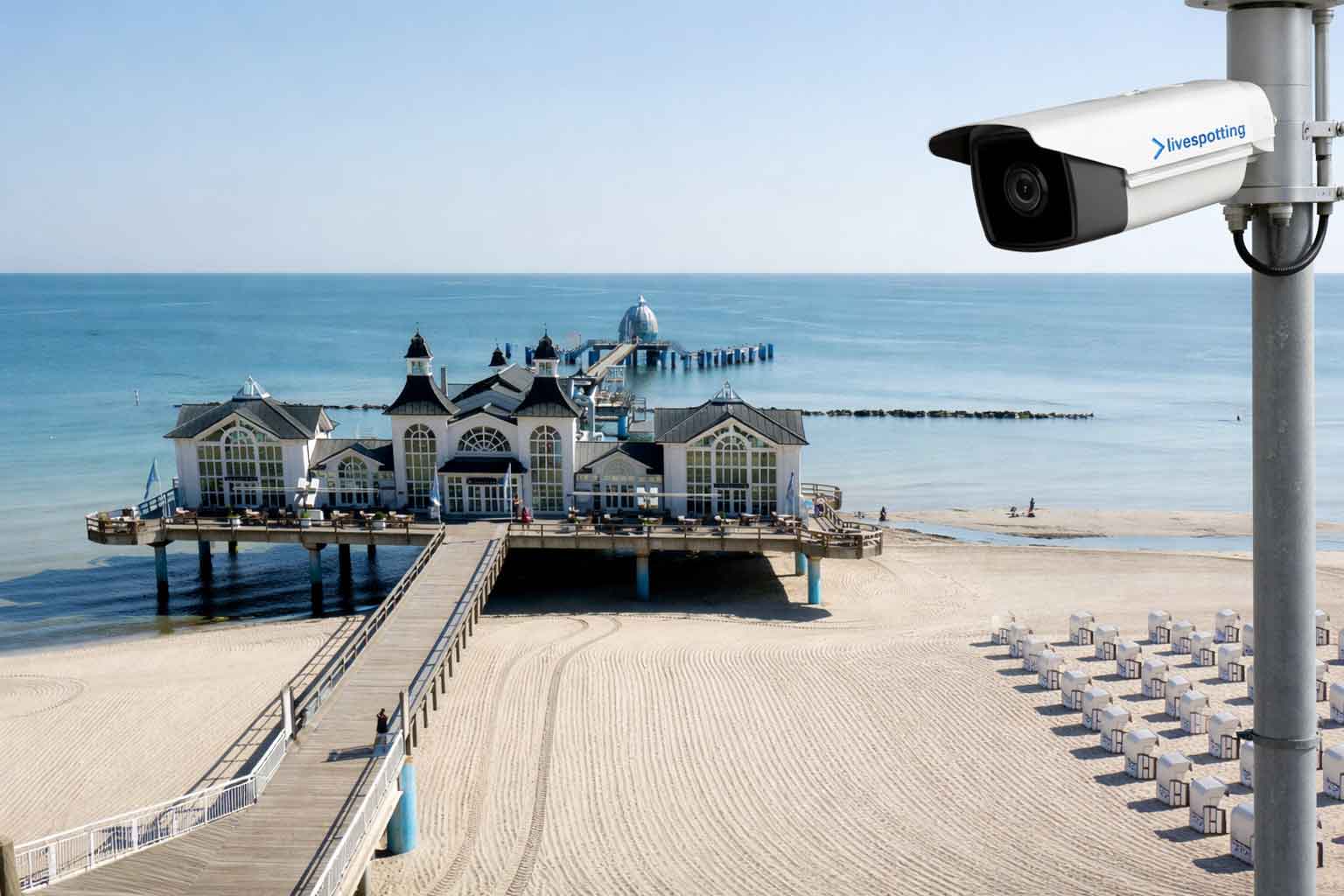 Eine weiße livespotting Bullet-Kamera, montiert an einem Mast, liefert einen Live-Stream von einem sonnigen Strand mit Seebrücke und Gebäude am Meer. Das Bild steht für effektives Tourismusmarketing durch Webcams an Küstendestinationen.
